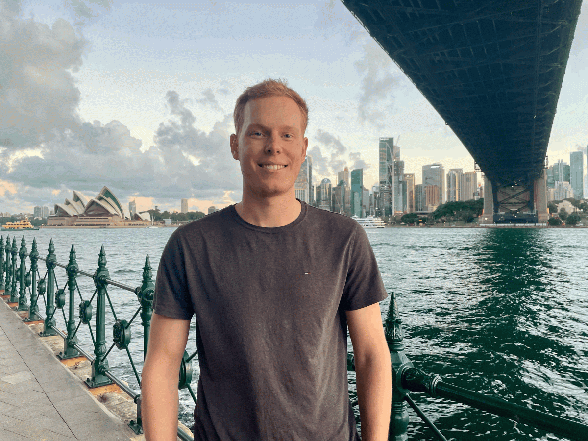 Kieran stands under the Sydney Harbour Bridge, with the Sydney Opera House in the background